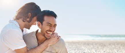 Zwei Männer lehnen sich an einander an am Strand mit Meereskulisse und hellem Sonnenlicht im Hintergrund