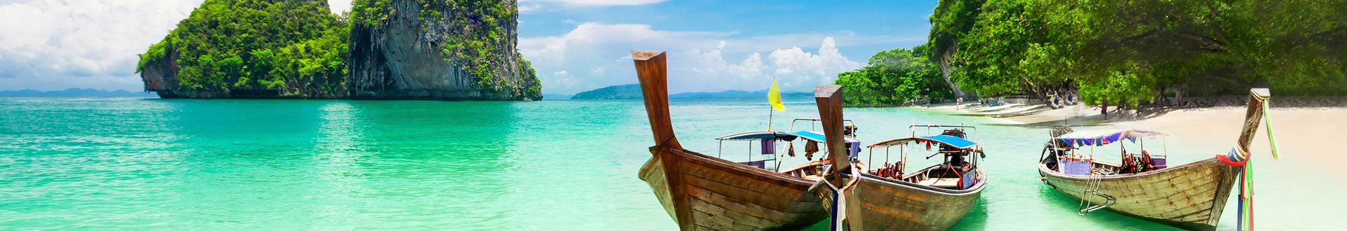 Drei traditionelle thailändische Longtail-Boote liegen am Strand vor einer tropischen Landschaft mit klarem Wasser und grünen Felsen.