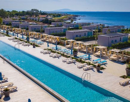 Luftaufnahme eines langgestreckten Pools mit Liegen und Sonnenschirmen, umgeben von modernen Campinghäusern nahe der Küste mit Blick auf das Meer.