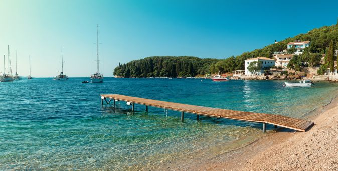 Holzsteg ragt in das klare Wasser einer Bucht am Kalami Beach auf Korfu, im Hintergrund Segelboote und bewaldete Hügel mit Häusern.