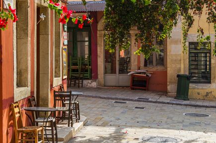 Sonniger Platz mit mehreren Holztischen und -stühlen vor einem Gebäude mit roten und gelben Wänden, darüber grüne Blätter und rote Blüten.
