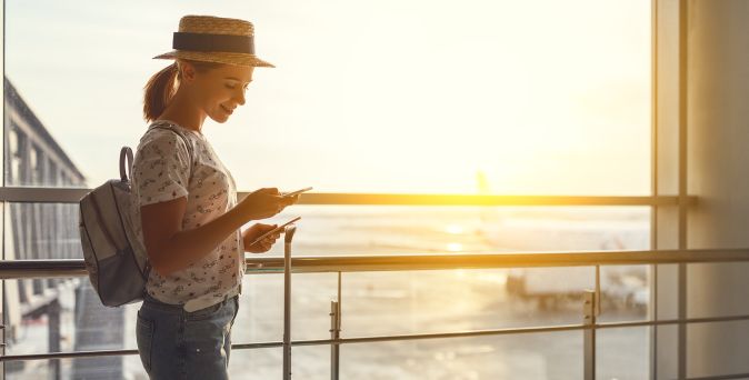 Junge Frau mit Hut und Rucksack am Flughafen, lächelt auf ihr Handy und hält ein Ticket in der Hand, während die Sonne durch das große Fenster scheint