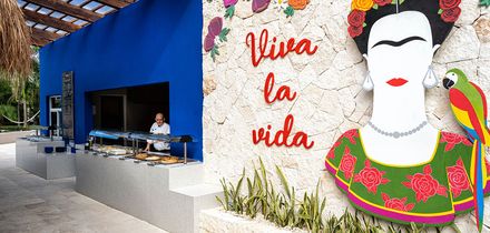 Wand mit farbiger Frida-Kahlo-Darstellung, Schriftzug 'viva la vida' und Papagei neben einem blauen Essensausgabefenster mit Person