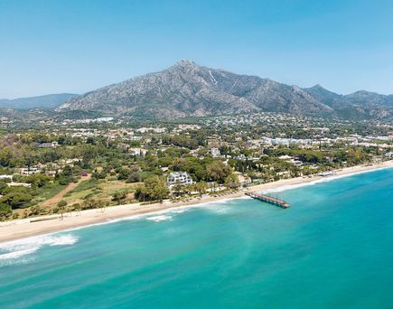 Spanische Küste mit Sandstrand, Pier und dahinter liegenden Gebäuden vor Bergkulisse unter klarem Himmel