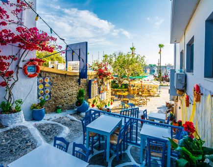 Gemütlicher Außenbereich eines griechischen Cafés mit blauen Tischen und Stühlen, bunten Blumen und Blick auf das Meer