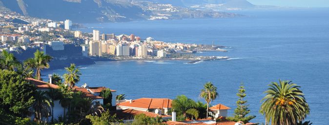 Blick auf eine Küstenstadt mit roten Dächern, Palmen und Bergen im Hintergrund am Meer