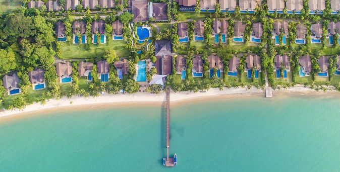 Luftaufnahme einer Strandresortanlage mit Reihen von Bungalows, privaten Pools, Palmen und einem Steg, der ins Meer führt