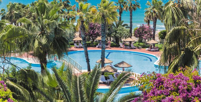 Hotelpool mit Sonnenschirmen und Liegestühlen umgeben von Palmen und blühenden Büschen mit Blick auf das Meer im Hintergrund