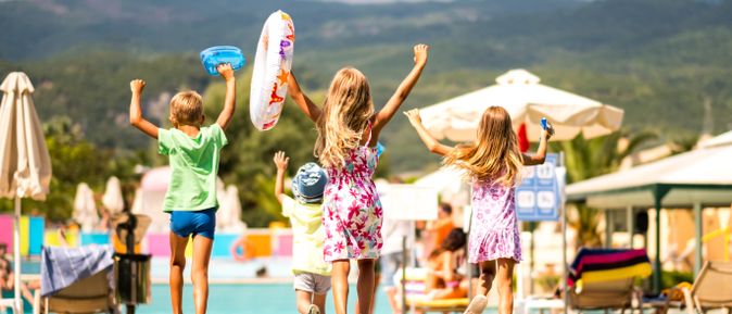 Vier Kinder laufen fröhlich mit Badeutensilien auf einen Hotelpool zu; im Hintergrund Sonnenschirme und Berge