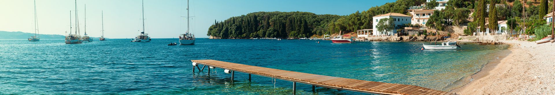 Strand mit klarem Wasser, Holzsteg und mehreren Segelbooten vor bewaldetem Küstenabschnitt