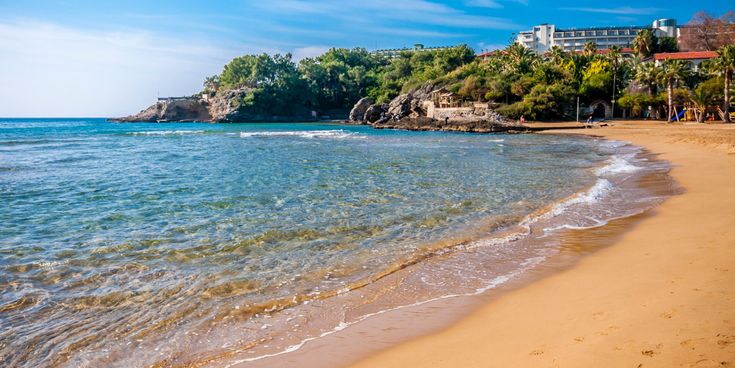 Goldener Sandstrand von Incekum an der türkischen Riviera mit sanften Wellen, klarem Wasser und grünen Palmen, im Hintergrund Hotels und Felsenlandschaft