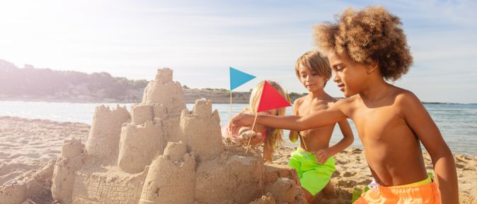Kinder bauen eine große Sandburg am Strand und stecken bunte Fahnen hinein, Meer im Hintergrund