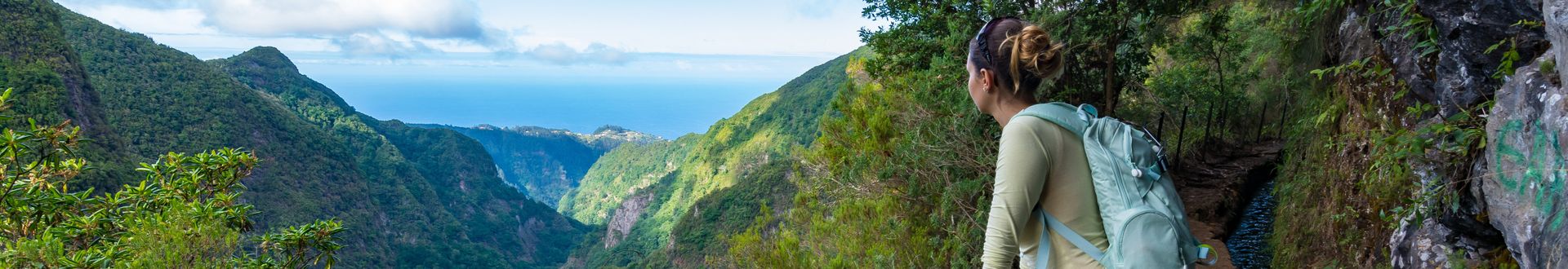 Person mit Rucksack auf der Levada do Alecrim auf Madeira mit Blick über ein grünes Tal, bewaldete Berghänge und den Atlantik am Horizont