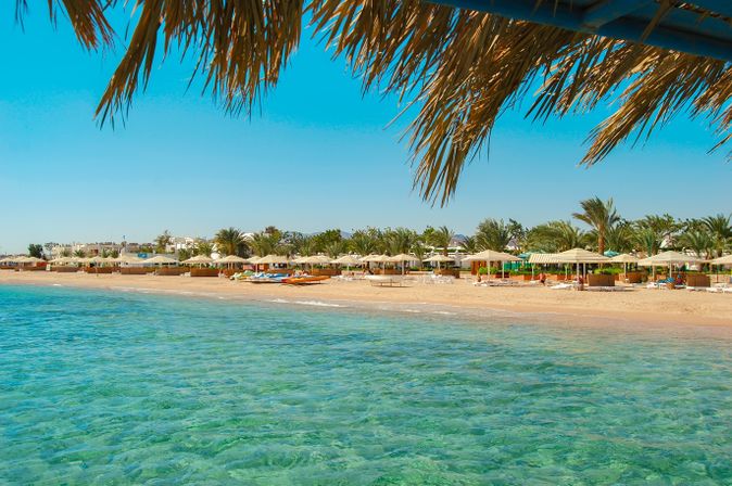 Klares türkisfarbenes Meer vor einem Sandstrand in Safaga mit Palmen und Sonnenschirmen unter blauem Himmel