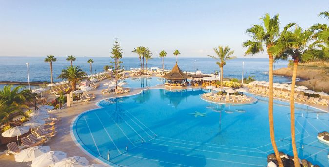 Großer, geschwungener Hotelpool mit zentraler Barinsel, umgeben von Liegestühlen und Palmen, mit Meerblick im Hintergrund.