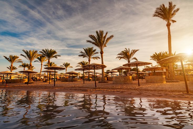 Strand mit Palmen und Sonnenschirmen in Hurghada bei Sonnenuntergang, der Sand und das ruhige Meer spiegeln warmes Licht wider