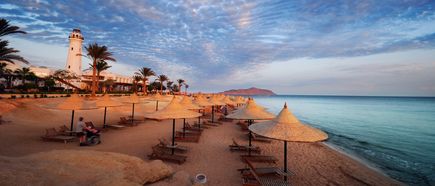 Strand mit Sonnenschirmen aus Stroh, Liegestühlen und einem Leuchtturm bei Sonnenuntergang in Sharm el-Sheikh