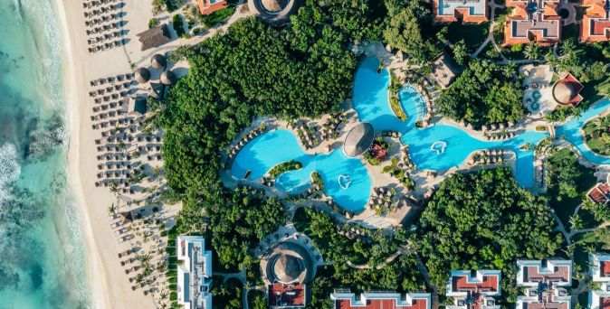 Luftaufnahme eines Resorts mit mehreren geschwungenen Pools, umgeben von grünen Bäumen, Gebäuden und einem Sandstrand mit Liegestühlen am Meer