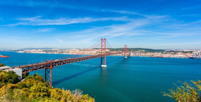 Rote Hängebrücke Ponte 25 de Abril über blauem Wasser mit Stadt Lissabon im Hintergrund unter blauem Himmel