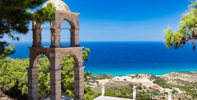 Steinerner Glockenturm mit drei Bögen vor blauem Meer und Küstenlandschaft mit Bäumen