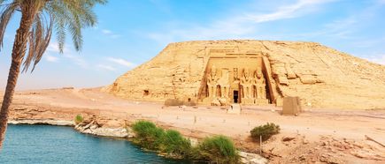 Felsentempel von Abu Simbel mit vier großen sitzenden Statuen am Eingang, Palmen und Wasser im Vordergrund