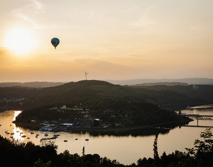 Landschaft mit See, Insel, Windrad auf Hügel und Heißluftballon im Abendhimmel