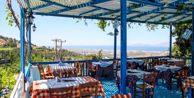 Überdachte Außenterrasse eines Restaurants mit blau-weißer Pergola, rot-weiß karierten Tischdecken und Holztischen mit Stühlen, im Hintergrund Blick auf grüne Hügel und das Meer.