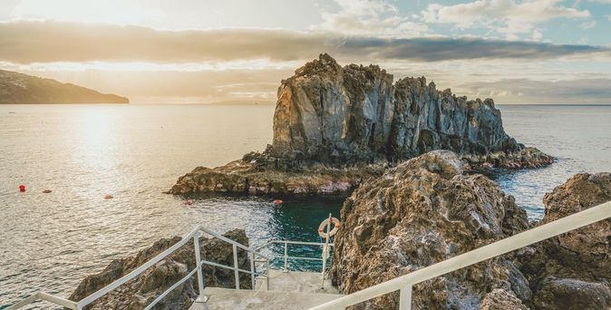 Felsinsel im Meer vor der Küste des Hotels Pestana Vila Lido Madeira bei Sonnenuntergang, Treppe und Geländer im Vordergrund