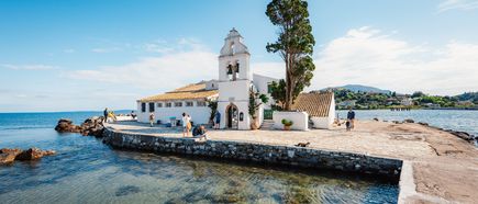 Klostergebäude mit Glockenturm auf einer kleinen Landzunge im Meer mit Spaziergängern und Bäumen