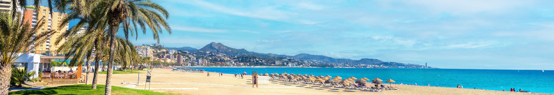 Blick auf einen Strand mit goldenem Sand und sanften Wellen, umgeben von hohen, gelben Felsen und blauem Himmel mit Wolken.
