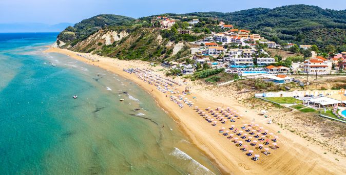 Luftaufnahme eines langen Sandstrands mit Reihen von Sonnenschirmen und Liegen, dahinter bewaldete Hügel und Gebäude.