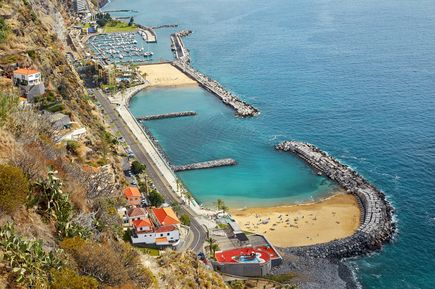 Luftaufnahme der Praia da Calheta auf Madeira mit goldfarbenem Sandstrand, türkisfarbenem Wasser, Hafenanlage, Wellenbrechern und Küstenstraße entlang der Steilküste