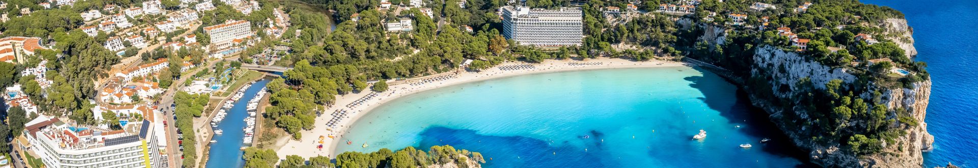 Luftaufnahme der Bucht Cala Galdana auf Menorca mit türkisblauem Wasser, Sandstrand mit Sonnenschirmen, Hotels und umliegendem Pinienwald