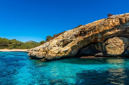 Cova des Pirata in Mallorca, eine große Felsformation mit einer Höhle, umgeben von klarem, blauem Wasser und grüner Vegetation im Hintergrund.