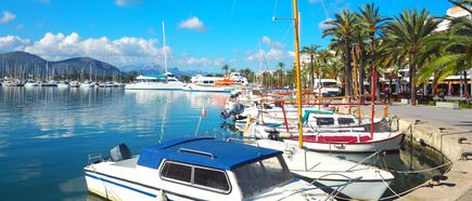Boote und Yachten im Hafen von Alcúdia auf Mallorca, gesäumt von Palmen und Restaurants, mit Bergen im Hintergrund