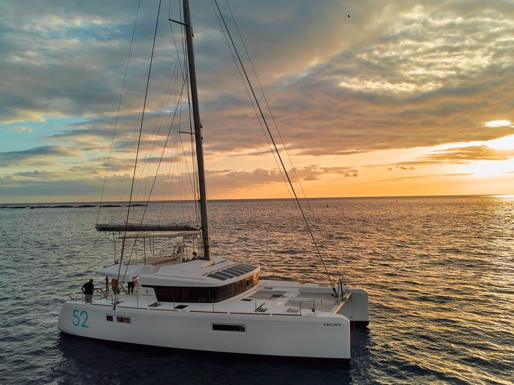 Weißer Katamaran segelt bei Sonnenuntergang auf dem Meer, der Himmel ist von Wolken und goldenem Licht geprägt
