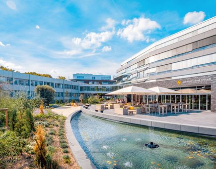 Firmengebäude mit Terrasse und Sitzbereichen neben einem Wasserlauf, blauer Himmel mit Wolken