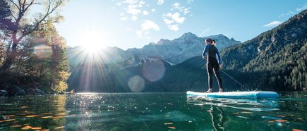 Person steht auf einem Stand-up-Paddle-Board auf einem klaren Bergsee im Sonnenlicht, umgeben von bewaldeten Hängen und Gipfeln