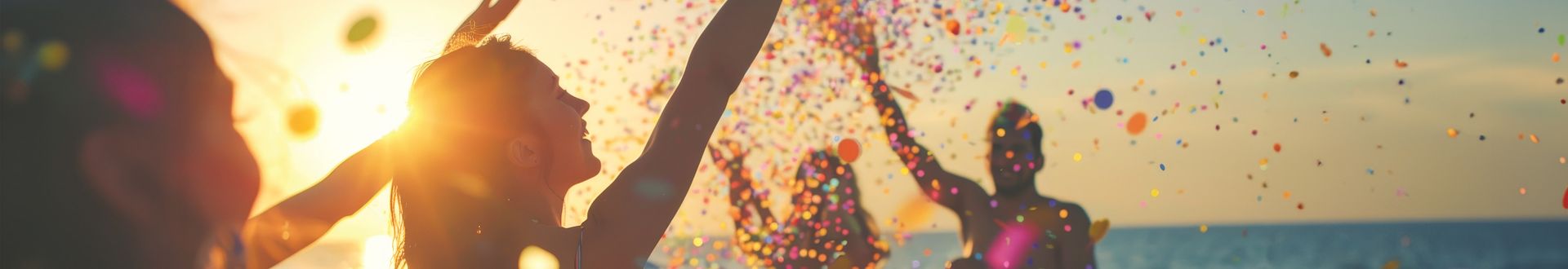 Mehrere Personen am Strand werfen buntes Konfetti in die Luft bei Sonnenuntergang über dem Meer.