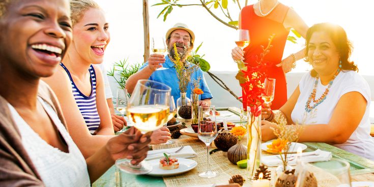 Gruppe fröhlicher Menschen sitzt an einem festlich gedeckten Tisch im Freien und stößt mit Weingläsern an