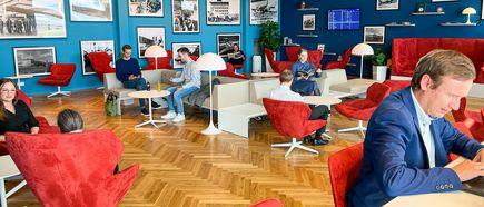 Moderne Lounge am Flughafen Stuttgart mit komfortablen Sitzbereichen, roten Sesseln, Arbeitsplätzen und Reisenden in entspannter Atmosphäre vor historischen Fotografien der Flughafengeschichte