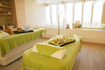Heller Massageraum mit zwei Liegen, grünem Überwurf und Tabletts mit Massageutensilien vor großen Fenstern mit Sitzbank