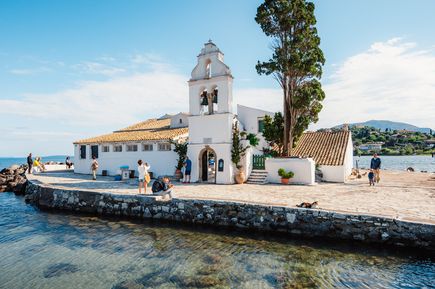 Klostergebäude mit Glockenturm auf einer kleinen Landzunge im Meer mit Spaziergängern und Bäumen