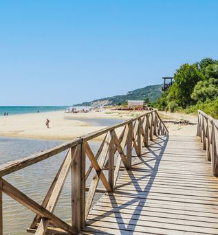 Holzsteg mit Geländer führt an einem Sandstrand entlang mit Meer und bewaldeten Hügeln im Hintergrund