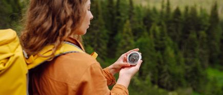 Eine Person mit gelbem Rucksack hält einen Kompass in den Händen und blickt in die Natur, Orientierung beim Wandern