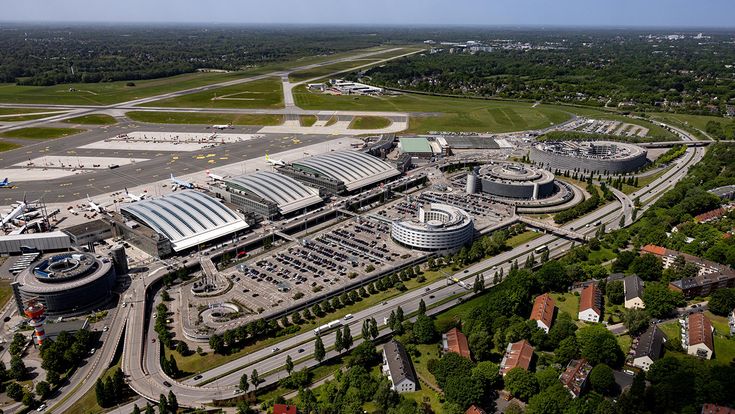 Weitläufige Luftaufnahme des Flughafens Hamburg mit Terminals, Vorfeld, Start- und Landebahnen sowie Parkhäusern, Straßenanbindung und umliegenden Wohngebieten im Grünen