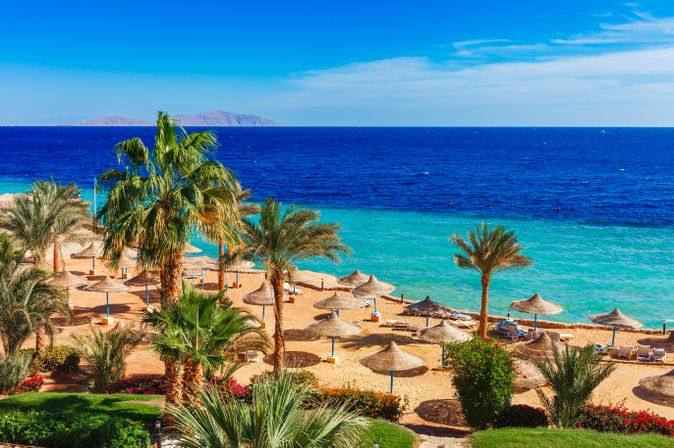 Strand mit Palmen und Sonnenschirmen am klaren Meer in Sharm el-Sheikh, Blick auf die Tiran-Insel am Horizont