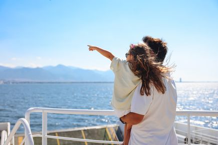 Kind auf dem Arm einer erwachsenen Person an Deck eines Schiffes mit Blick über das Meer und die Küste, während das Kind auf den Horizont zeigt