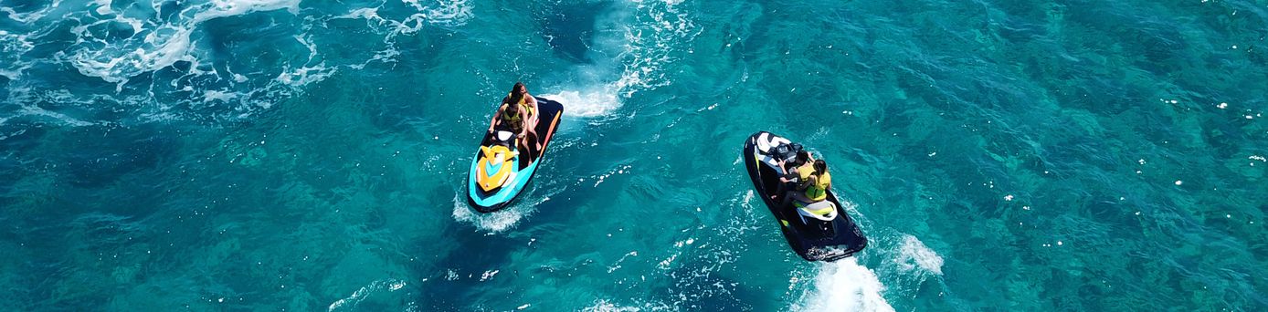 Zwei Jetskis mit jeweils einer Person fahren auf blauem Wasser und hinterlassen weiße Wasserstrahlen.
