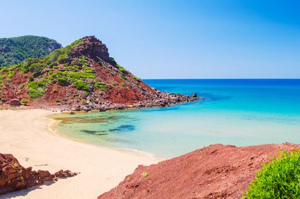 Blick auf die Bucht Cala del Pilar auf Menorca mit roter Felsküste, goldenem Sandstrand und türkisblauem Meer unter wolkenlosem Himmel